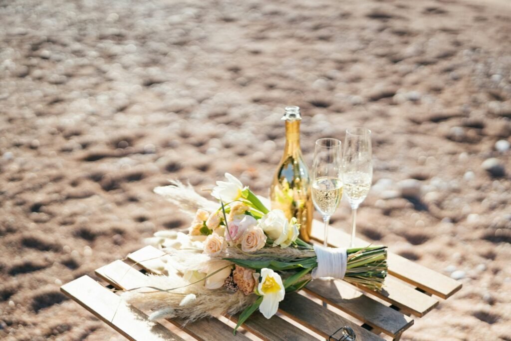 Ein Hochzeitsstrauß aus Blumen liegt auf einem Holztisch neben einer Flasche Champagner am Strand